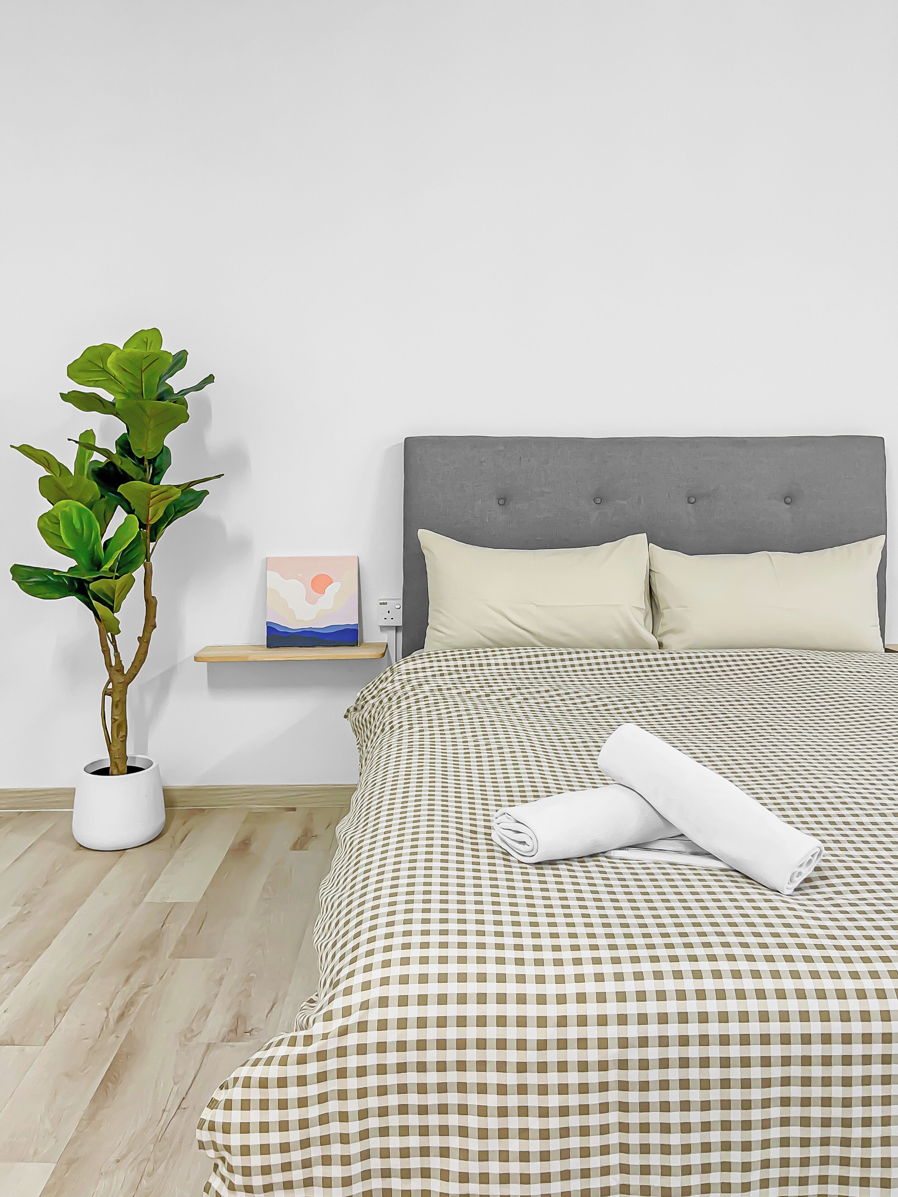 Una cama bien hecha con una colcha a cuadros, un cabecero tapizado de color gris y una almohada blanca. Un toalla blanca enrollada está colocada sobre la cama.