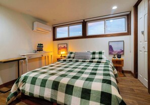 A bedroom with a checkered bedspread, a nightstand with a lamp, a window with a view, and an air conditioning unit.