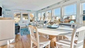 Ein heller, geräumiger Wohnbereich mit großem Fenster zum Strand, einem weißen Esstisch mit Stühlen und einer gemütlichen Sitzecke mit Sofa und Sessel.