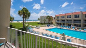A hotel with a swimming pool, a beach view, and a fenced balcony area.