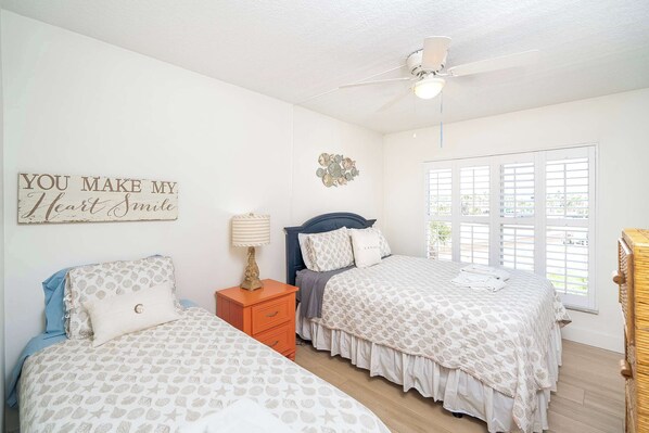 Quarto com duas camas, ventoinha no teto, janela com persianas e um objeto decorativo na parede.