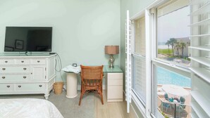 Un dormitorio con cama, cómoda, televisor, escritorio con silla y ventana con vista a una piscina y mobiliario exterior.
