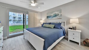 A bedroom with a bed, a ceiling fan, a nightstand, a lamp, a painting, and a view of a building through a glass door. - Pier Point South Ocean View Condo D-44 (St. Augustine)