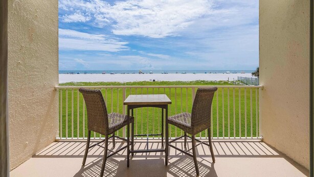 Een balkon met twee rieten stoelen en een kleine tafel, met uitzicht op een strand met een gazon en een hek.