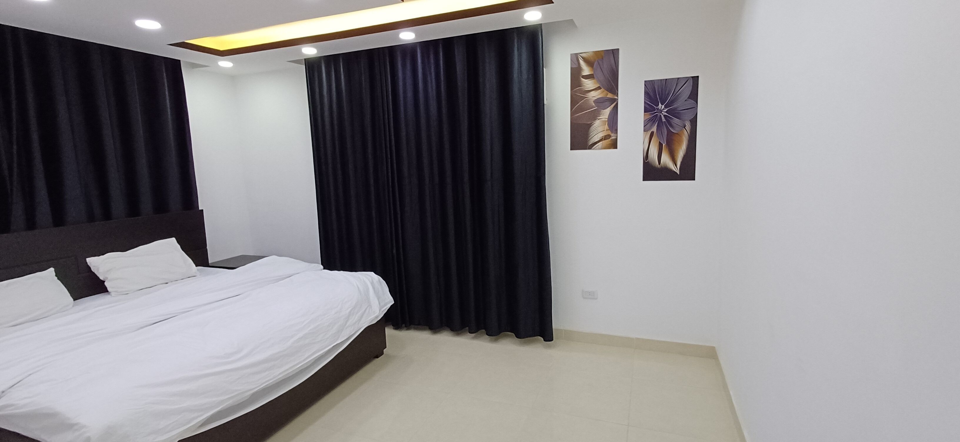 Un dormitorio moderno con una cama grande, cortinas oscuras y dos cuadros de flores colgados en la pared.