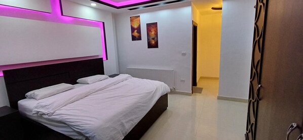 A modern bedroom with a large bed, white bedding, and a headboard. There are two wall-mounted paintings and a door leading to another room. - Laperla luxury apartments  (Amman)