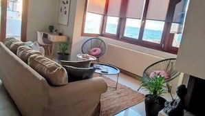 A living room with a sofa, a small table, and a chair. There is a window with a view of the sea. - Nova Sea View Apartment Seafront (Kokkini Hani)