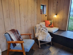 A cozy room with wooden walls, a blue chair, a cushioned bench, a small shelf with books, and a window with a view.