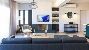 A modern living room with a large dark sofa, a TV mounted on the wall, and a ceiling fan. - Dione Seaside Apartment with Sea View (Anissaras)