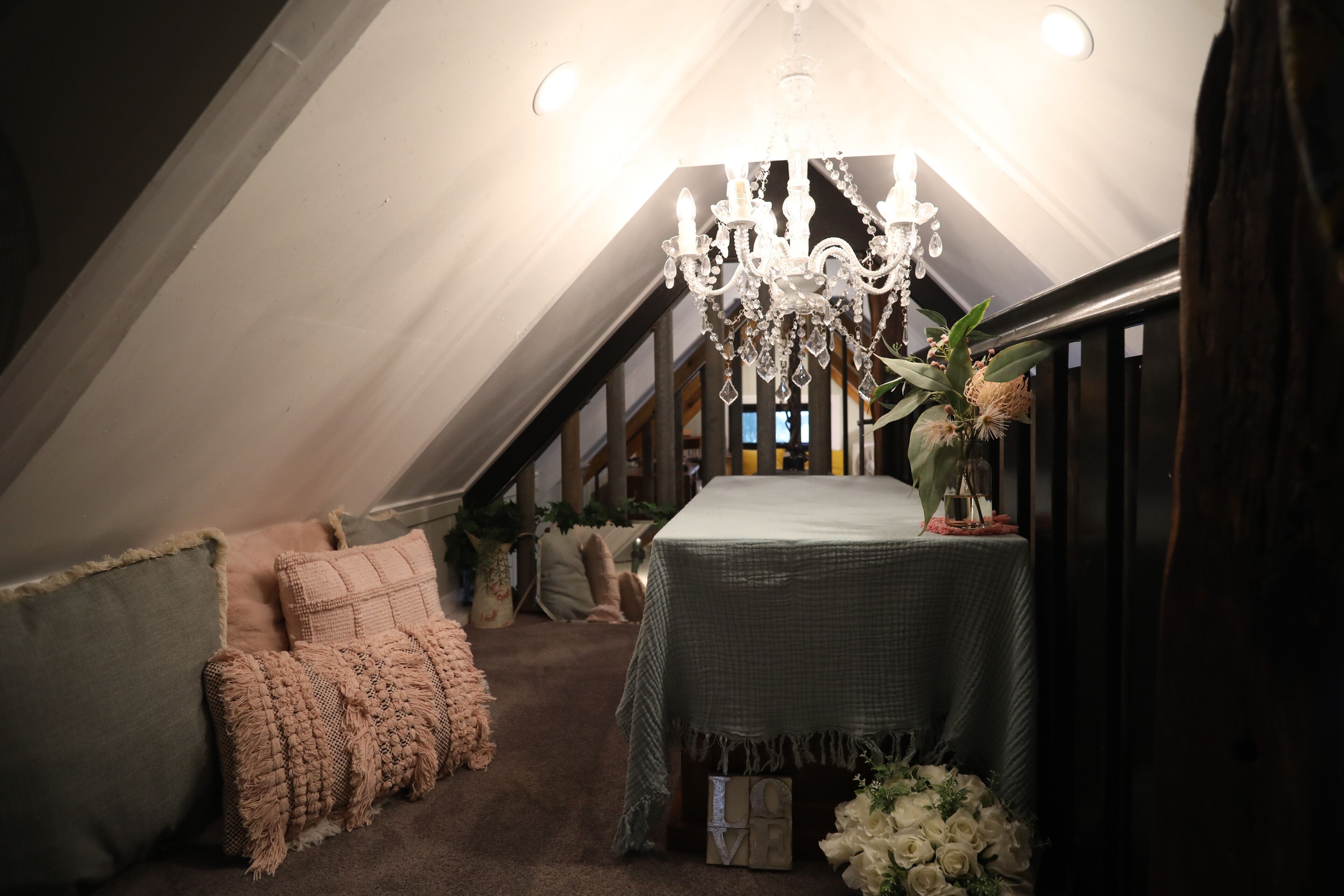 A cozy attic bedroom with a chandelier, a bed with a green bedspread, and a small table with a vase of flowers.