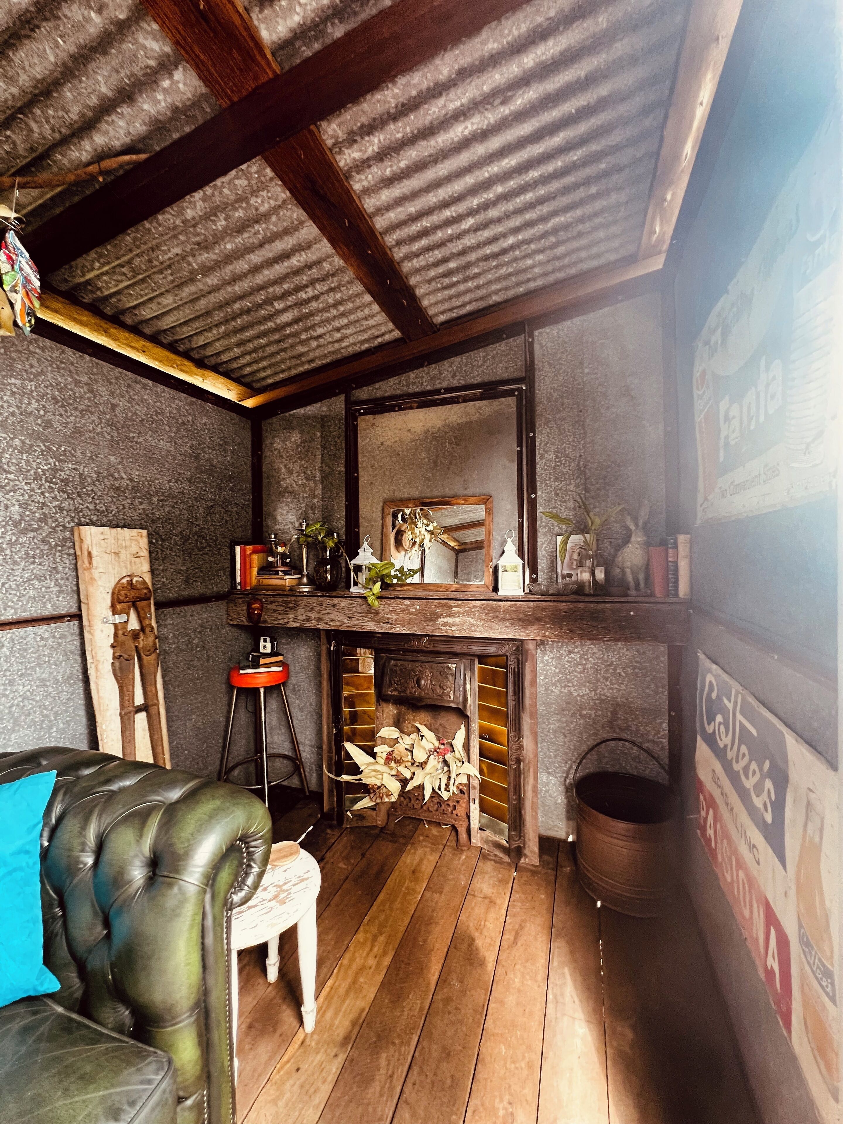 A rustic room with a wooden floor, a leather chair, a small stool, a fireplace with logs, and a mirror above it.