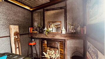 A rustic room with a wooden floor, a leather chair, a small stool, a fireplace with logs, and a mirror above it.