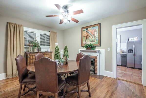 A dining room with a wooden table, four chairs, a ceiling fan, a fireplace, and a kitchen visible through an open door.