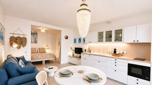A modern kitchen with white cabinets, a dining table with chairs, a chandelier, and a blue sofa.