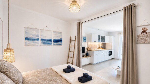 A bedroom with a bed, a bedside table, a lamp, and a view into the kitchen.
