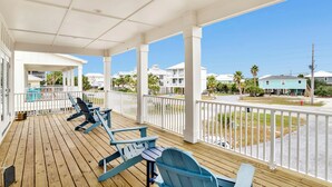 Un porche techado con sillas Adirondack azules, barandal blanco y vista a una zona residencial con casas y palmeras.