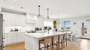 Una cocina moderna con armarios blancos, una isla grande con taburetes y lámparas colgantes, y vistas a través del ventanal.