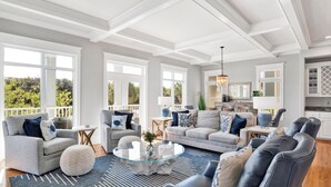 A spacious living room with a large glass coffee table, multiple seating options including sofas and chairs, and large windows offering a view of greenery outside. - Kiva Dunes Luxury Retreat | Private Pool | Gulf Views (Gulf Shores)