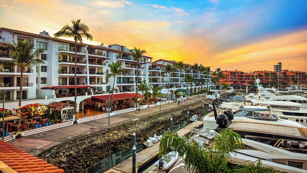 A marina with boats docked, a waterfront promenade with restaurants, and multi-story buildings with balconies.