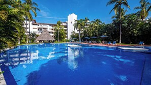 A swimming pool with lounge chairs and umbrellas, surrounded by palm trees and a building with a thatched roof.