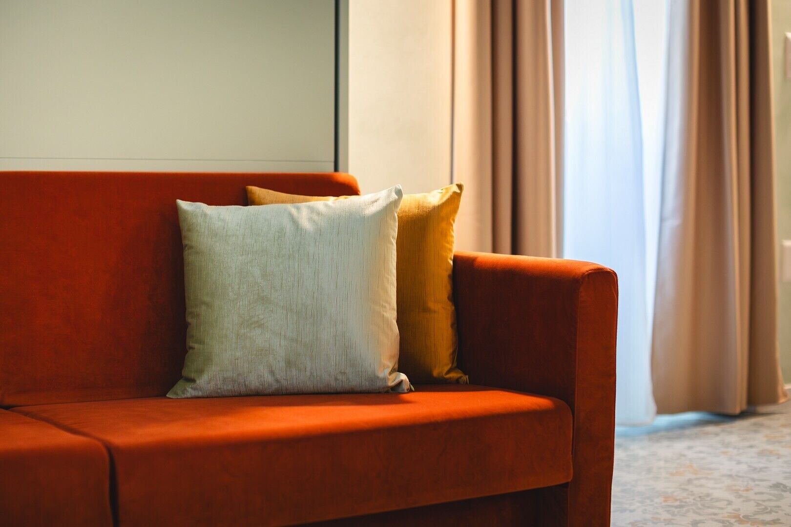 A red sofa with two pillows, a plain wall, and a window with curtains.