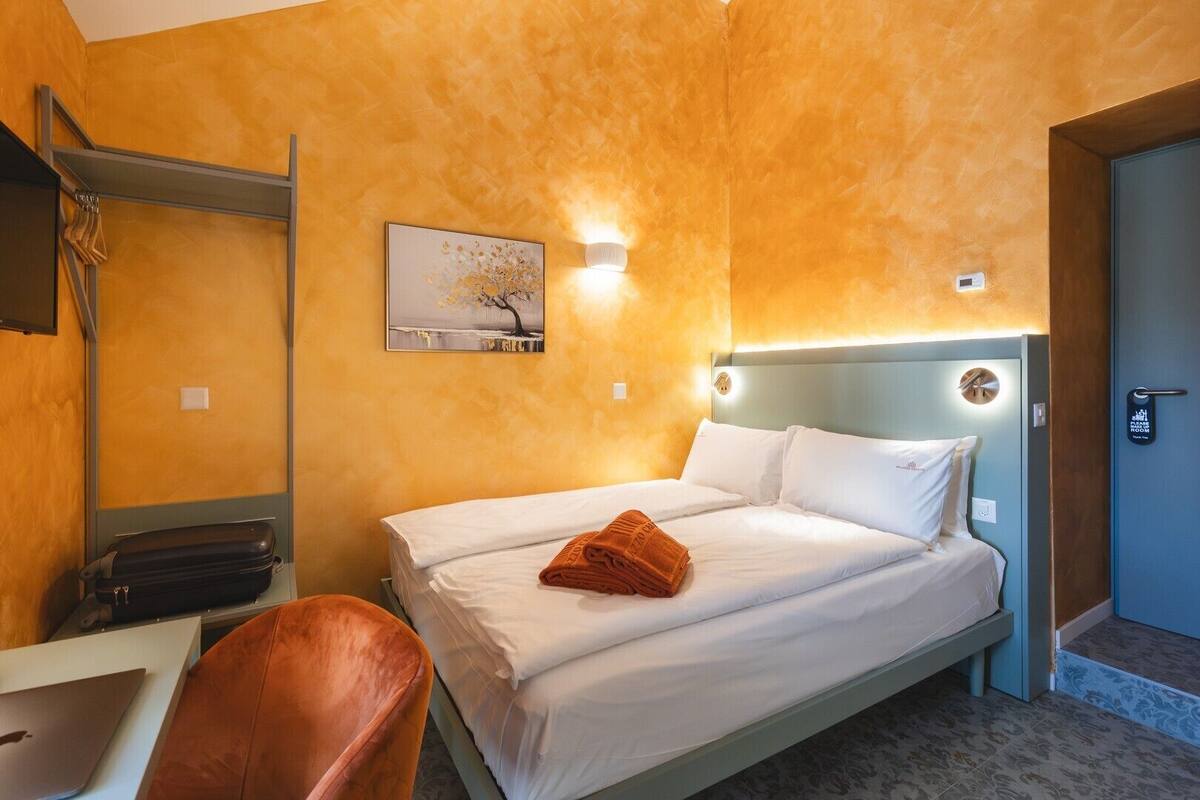 A hotel room with a bed, a desk, and a chair. There is a painting on the wall and a door with a handle.
