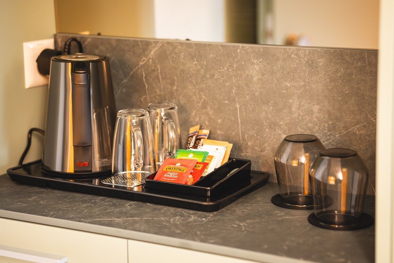 A kitchen counter with a coffee maker, four glass tumblers, a tray with Twinings English Breakfast tea bags, and two glass carafes.