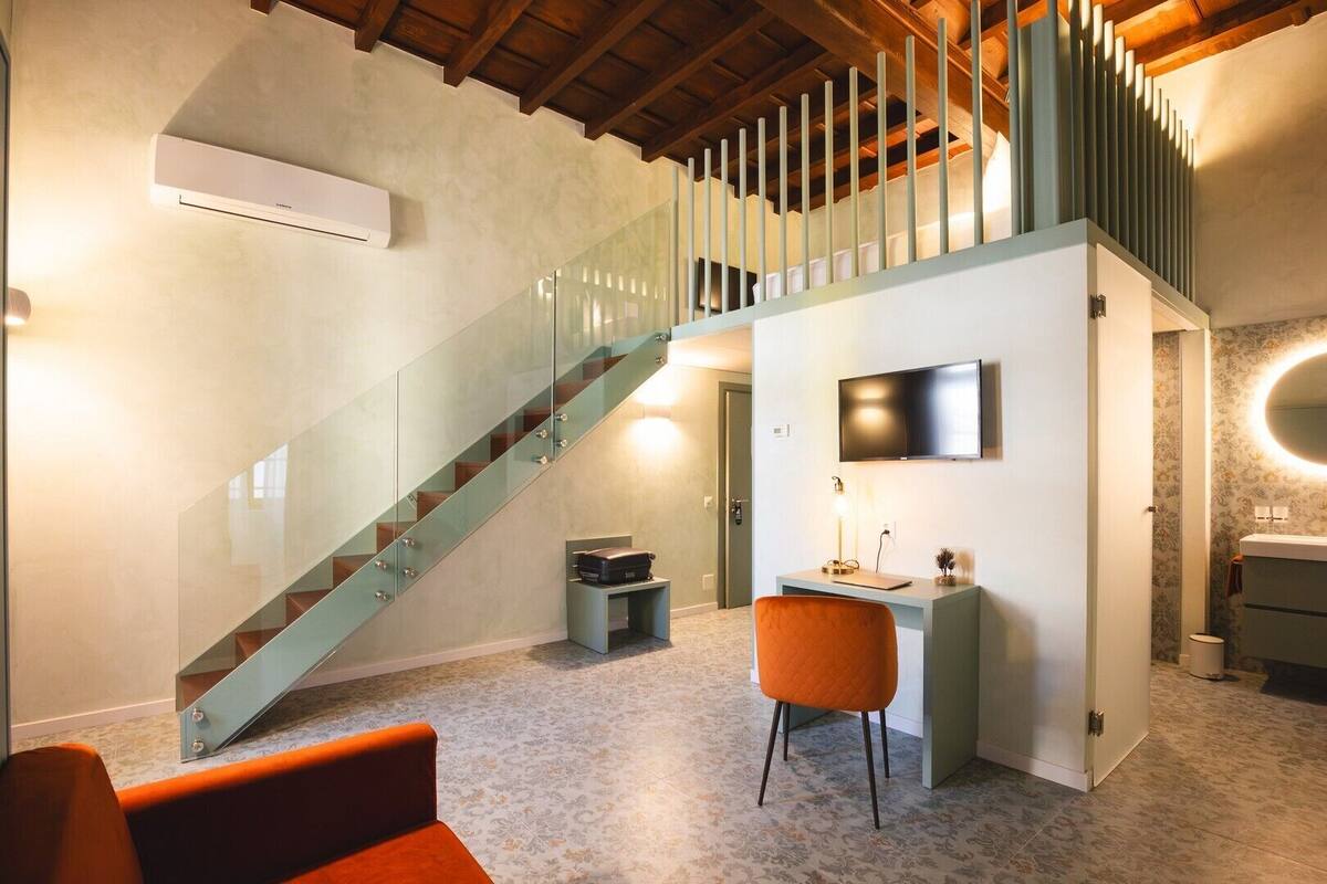 A modern interior with a glass staircase, a desk with a chair, a flat-screen TV, and a bathroom with a mirror.