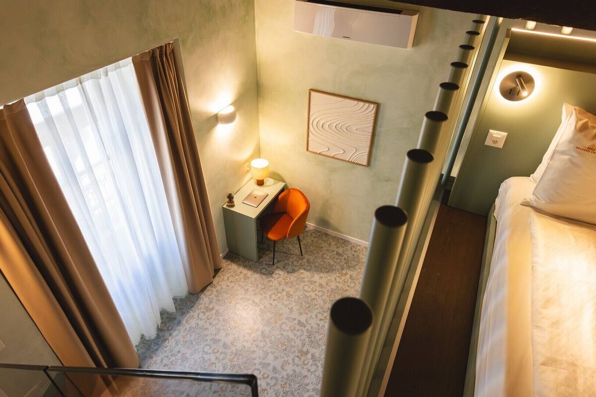 A compact hotel room with a bed, a desk with a laptop, a chair, and a small table with a lamp.