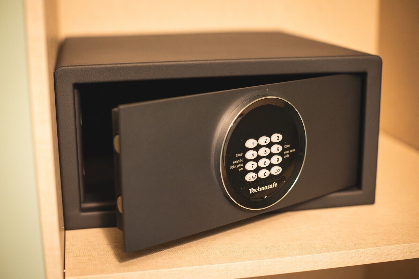 Ein schwarzer Tresor mit Tastatur, beschriftet mit 'Technosafe', auf einer Holzwandregal.