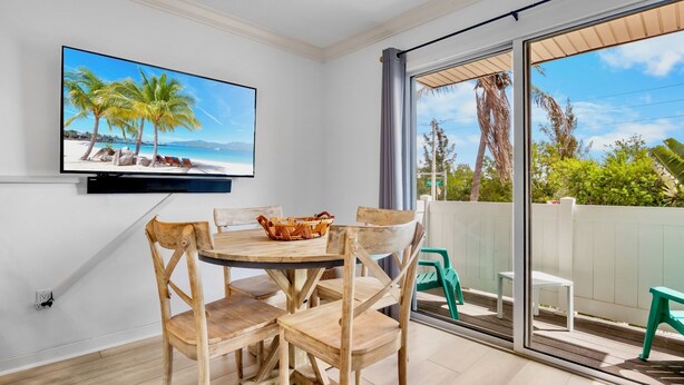 A room with a TV displaying a beach scene, a wooden dining table with chairs, and a sliding glass door leading to an outdoor area with chairs.