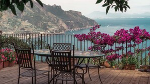 Una terraza de madera con una mesa y sillas, con vistas a un paisaje costero con montañas y el mar.