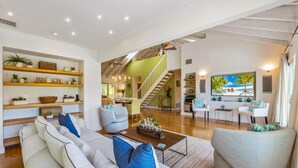 Amplia sala de estar con un sofá grande, una mesa de centro y un televisor. Se observa una escalera que conduce a un nivel superior, y estantes con plantas y objetos decorativos.