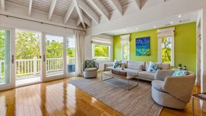 Un salón espacioso con piso de madera, un ventanal con vistas a la vegetación, una pared de acento verde y un techo con vigas a la vista.