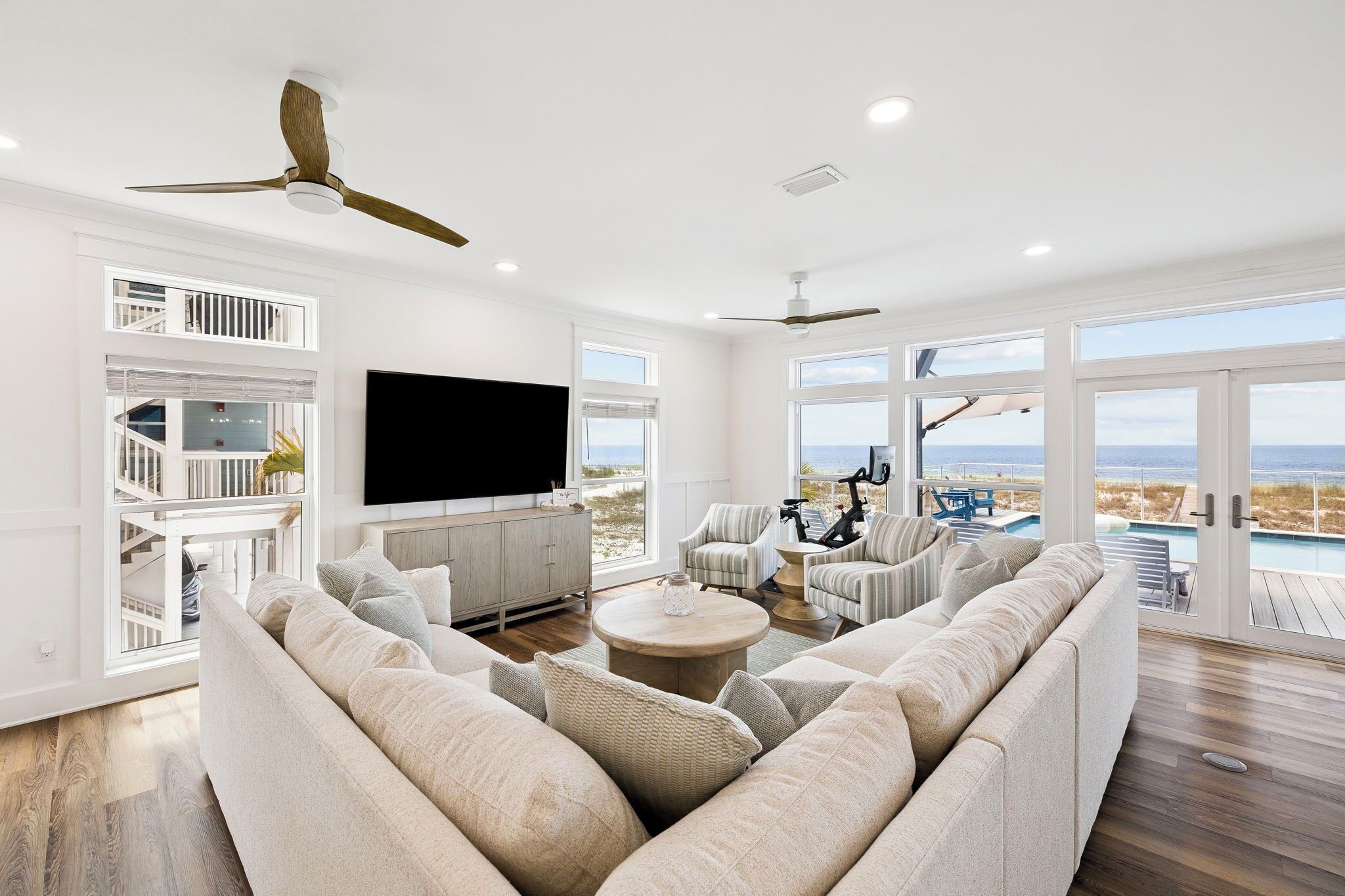 A spacious living room with a large sofa, a coffee table, and a TV. There are two ceiling fans, a balcony with a view, and a pool area visible through large windows.
