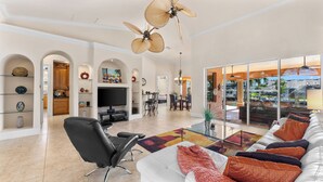 A spacious living room with a large sofa, black leather chair, and a glass coffee table. There are ceiling fans, a flat-screen TV, and a dining area with a table and chairs.