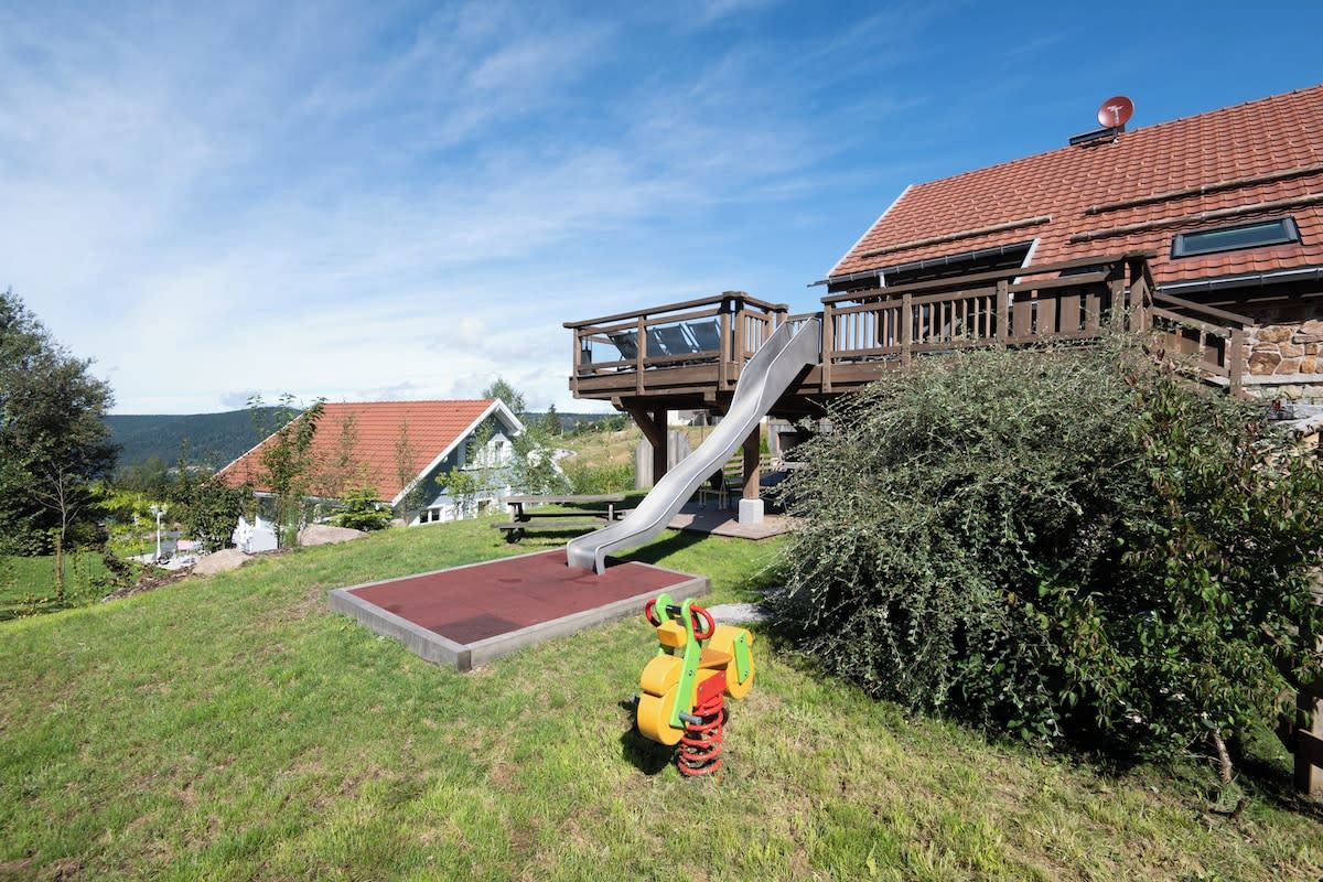 Un parque infantil con un tobogán y una estructura para escalar, rodeado de césped y un edificio con techo de tejas rojas.