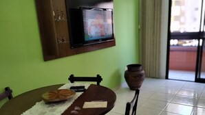 Un coin repas comprenant une table ronde en bois, quatre chaises, un téléviseur fixé sur un meuble en bois et une grande fenêtre donnant sur des immeubles.