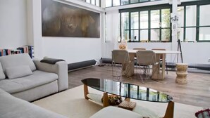 A spacious living area with a modern sofa, a glass coffee table, and a dining table with chairs. Large windows allow natural light to fill the room.
