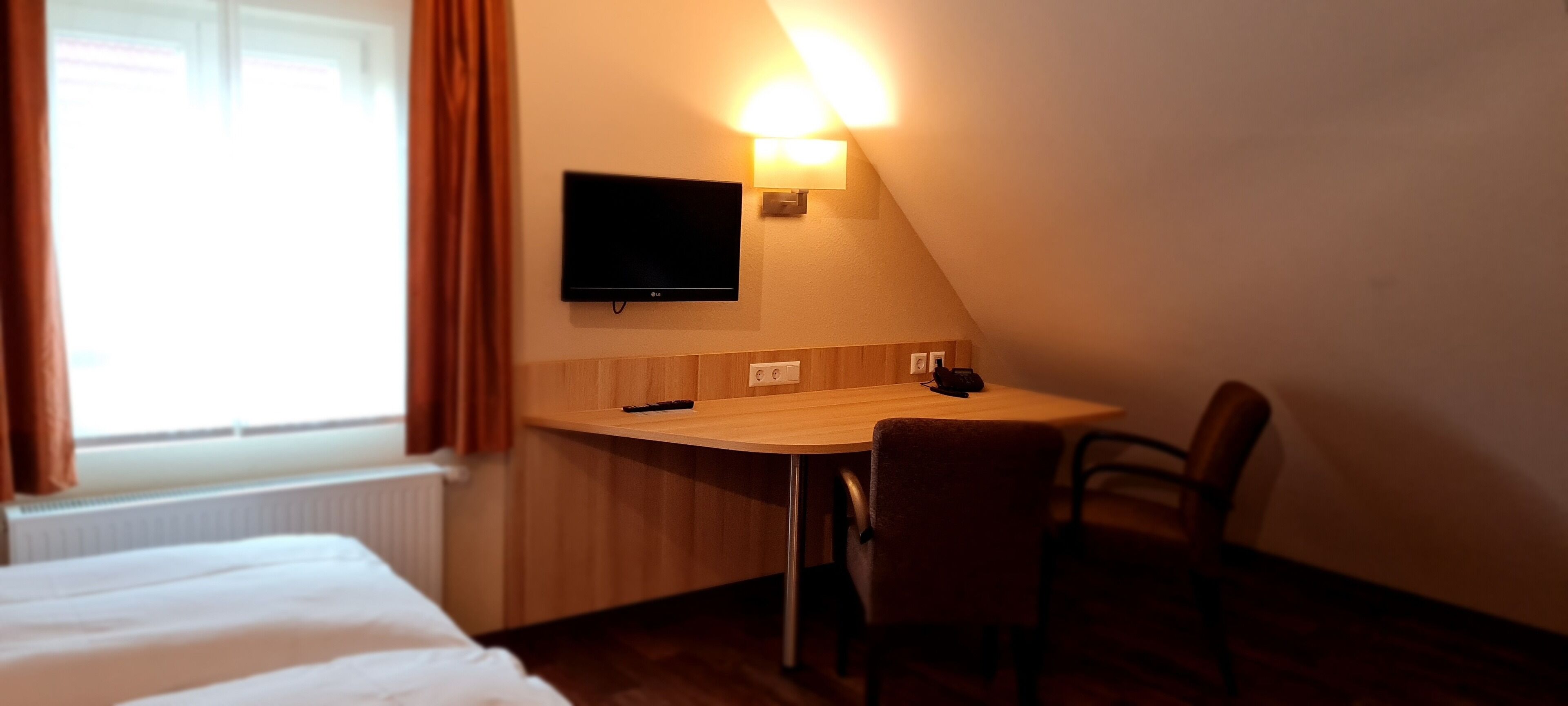 A hotel room with a bed, a desk with a chair, a TV mounted on the wall, and a window with curtains.