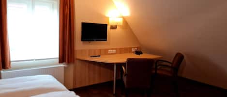 Habitación de hotel con cama, escritorio con silla, televisor montado en la pared y ventana con cortinas.