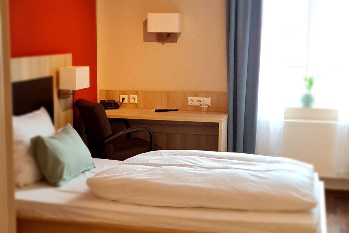 A bedroom with a wooden bed, a desk, and a chair. There is a window with curtains, a wall-mounted light, and a ceiling light.
