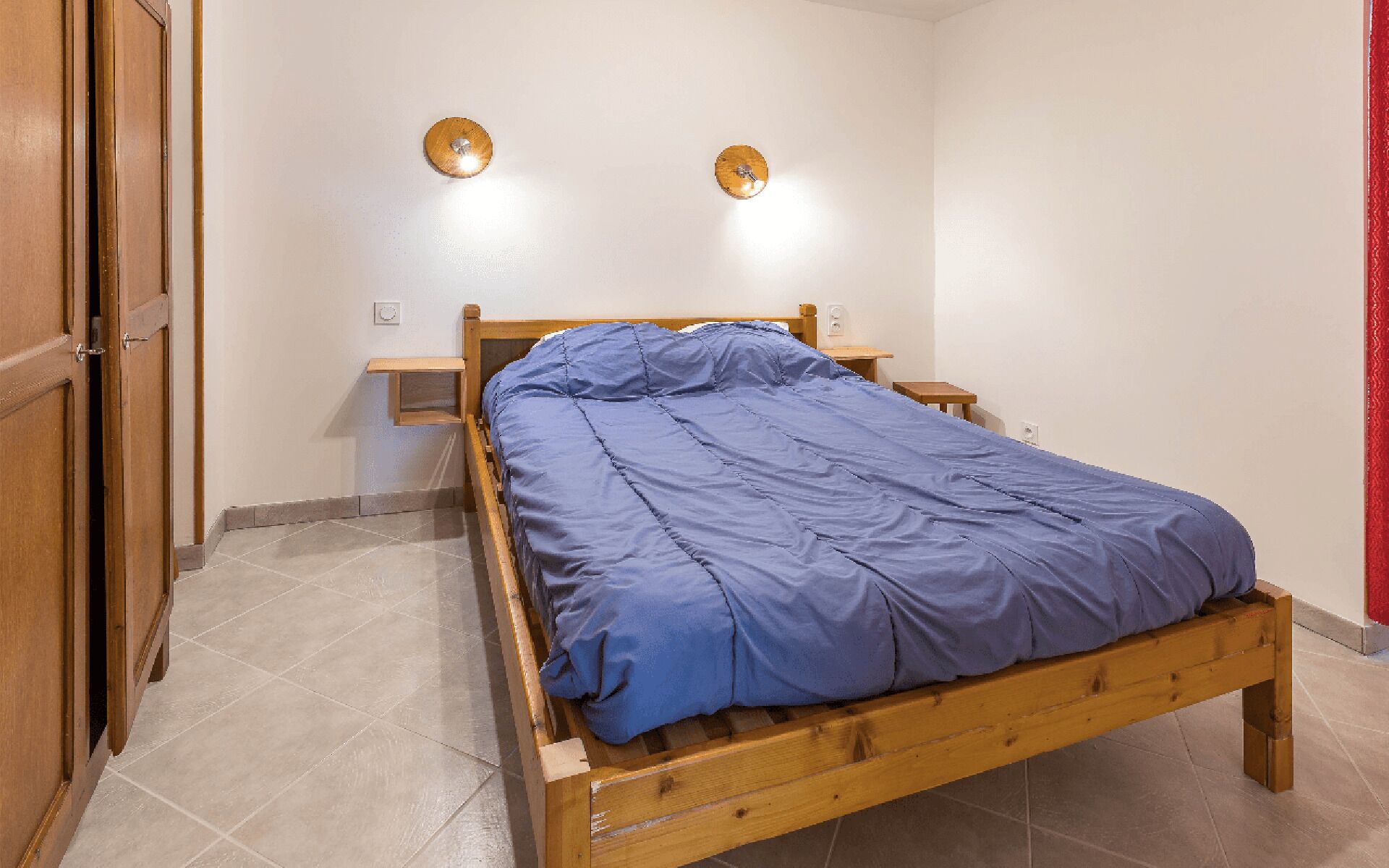 A single bed with a wooden frame and a blue mattress in a room with white walls and a tiled floor.