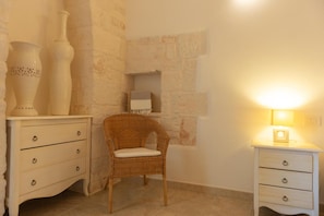 A room with a wicker chair, a white dresser with drawers, a small shelf with a box, and a wall-mounted light. - Villa in Itria Valley with Infinity Pool (Cisternino)