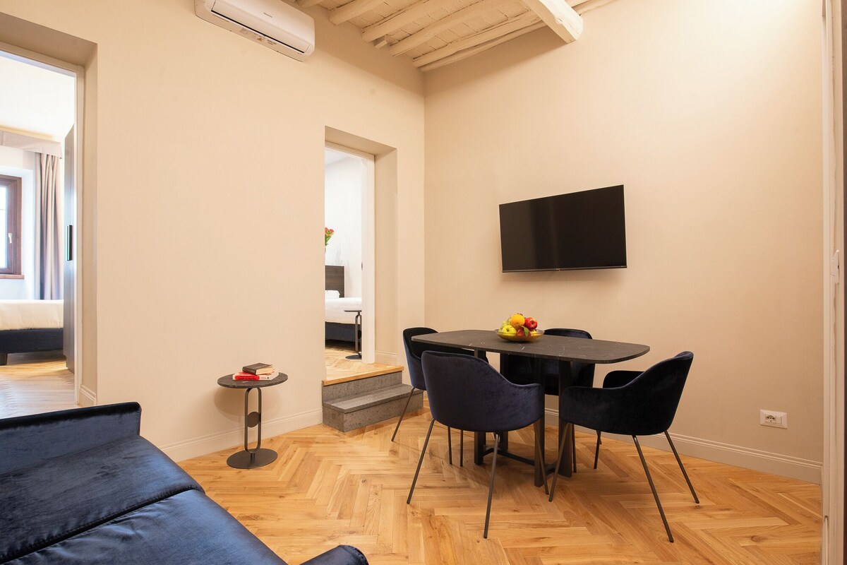 A modern living room with a sofa, a dining table with chairs, a TV mounted on the wall, and a small side table with a book and a bowl of fruit.