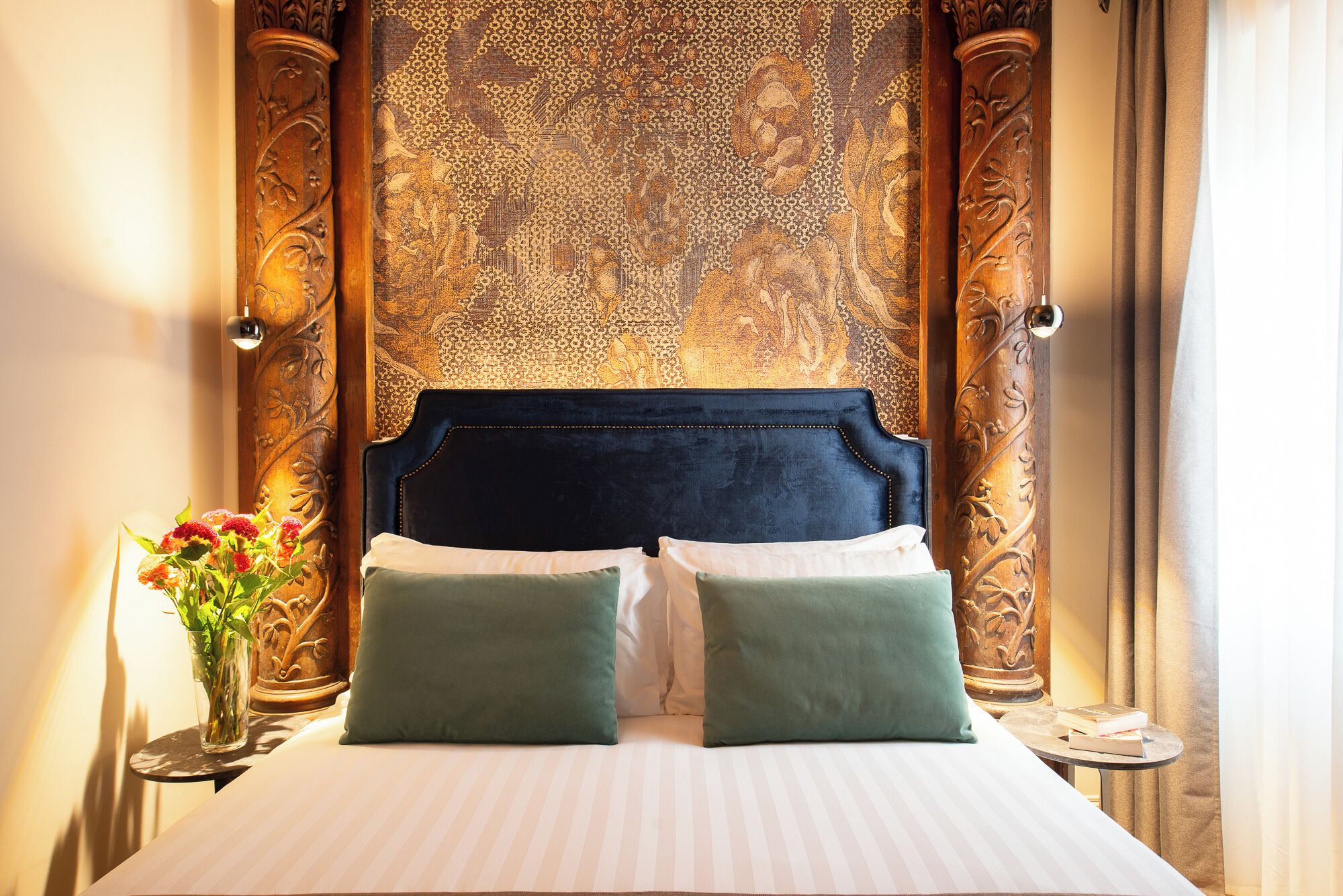 Een hotelkamer met een groot bed, twee kussens, een vaas met bloemen en een sierlijk hoofdeinde.