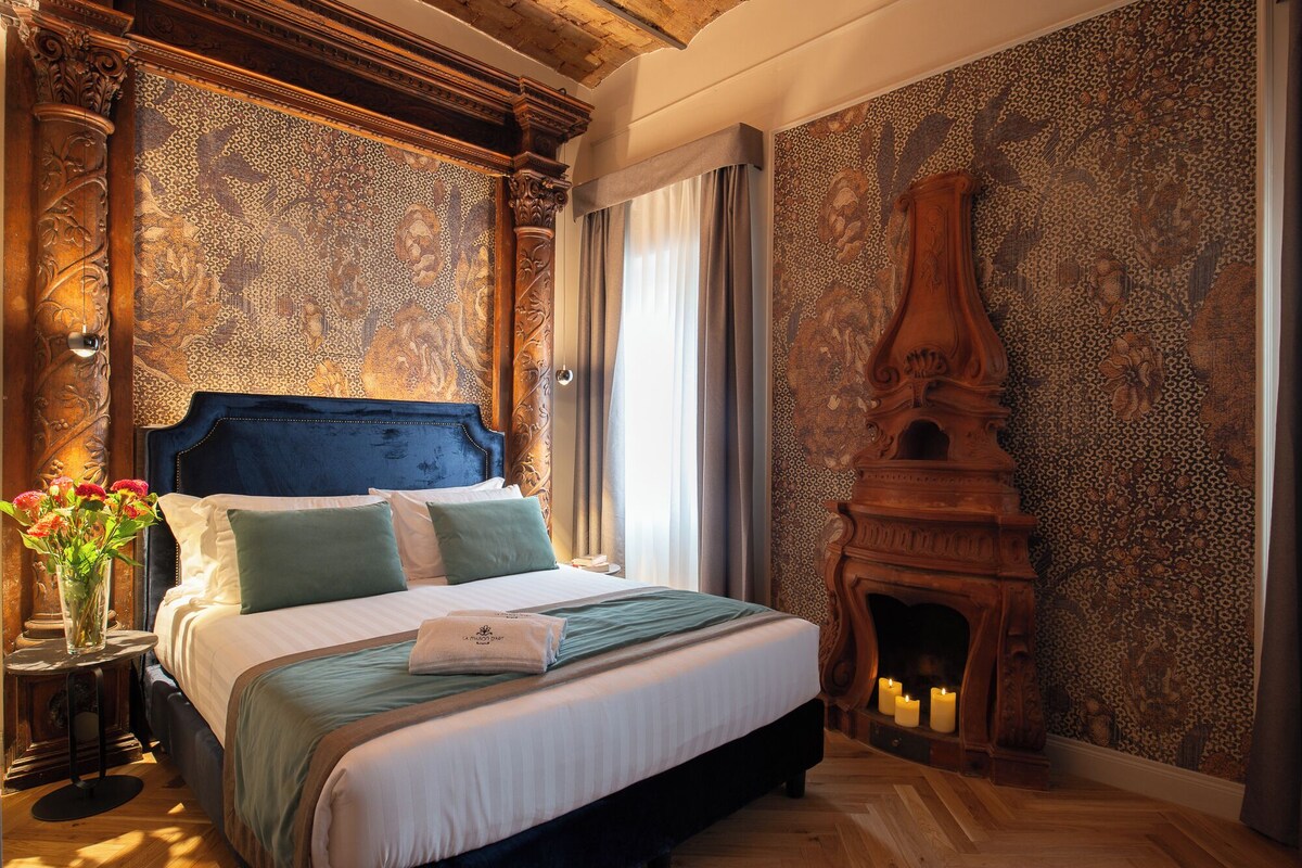 A bedroom with a large bed, ornate wooden headboard, a fireplace, and patterned wallpaper.