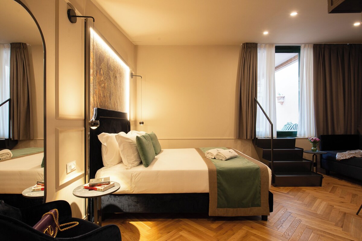 A hotel room with a bed, a small table with a book, a chair, a sofa, a staircase, and a large mirror.