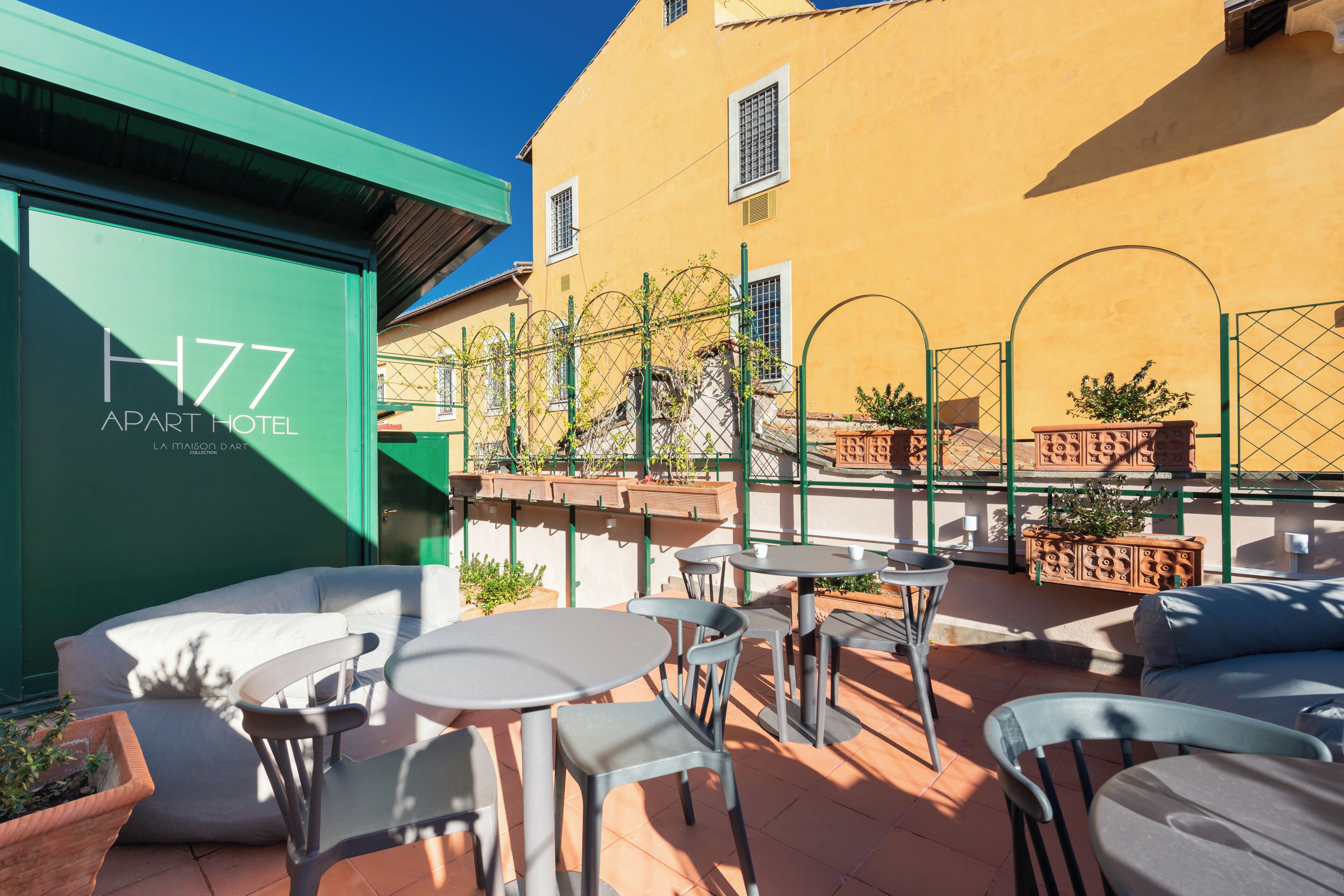 Une terrasse extérieure avec des tables et des chaises rondes, un bâtiment vert portant l’inscription « H77 APART HOTEL », et un bâtiment jaune en arrière-plan.
