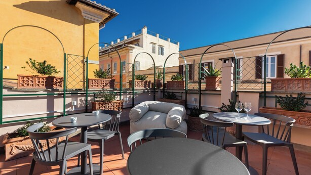 Une terrasse extérieure avec des tables et des chaises rondes, des plantes en pot et un bâtiment jaune en arrière-plan.